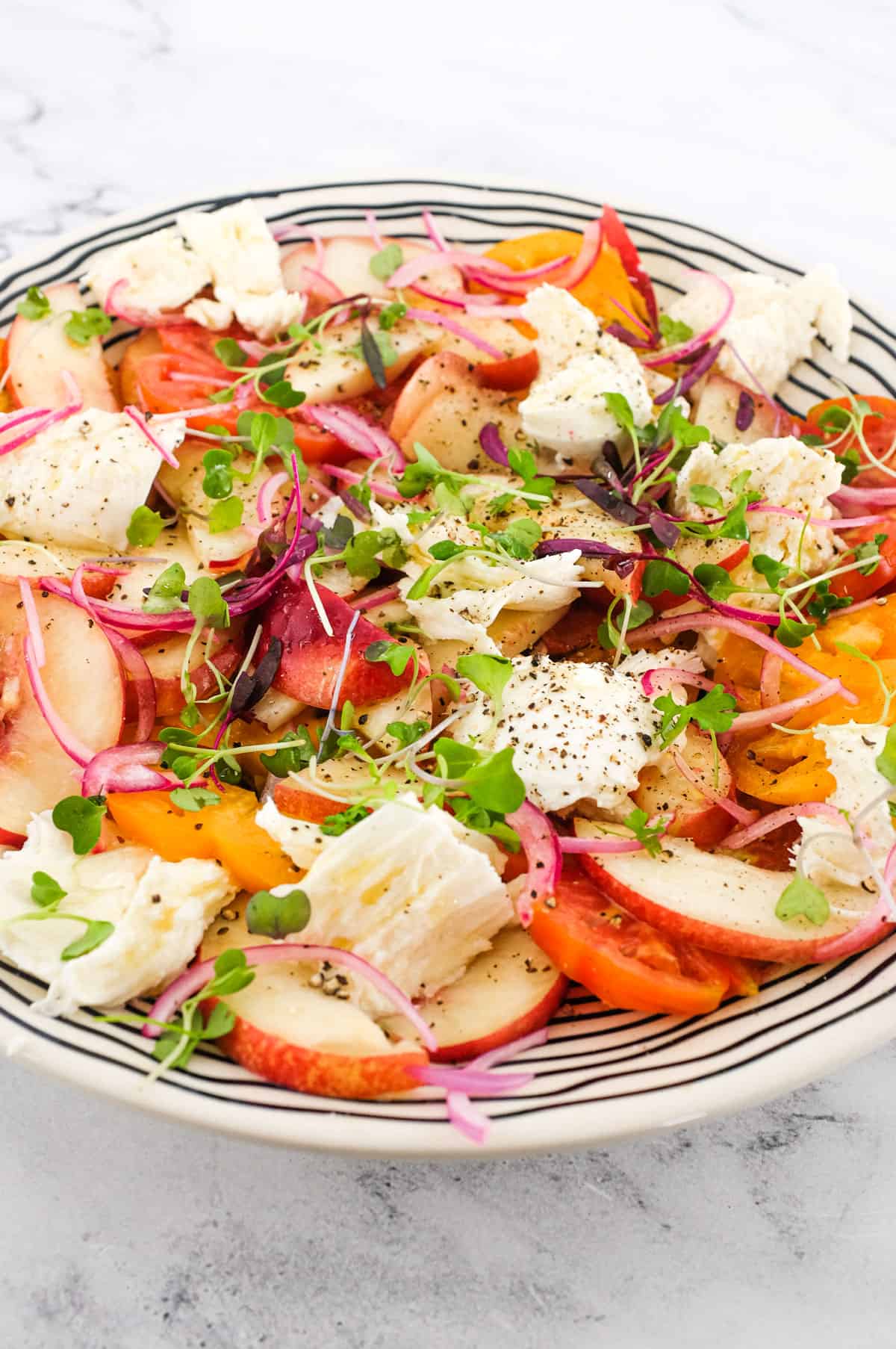 A salad of peaches, burrata cheese, tomato, red onion and micro greens sits on a marble countertop