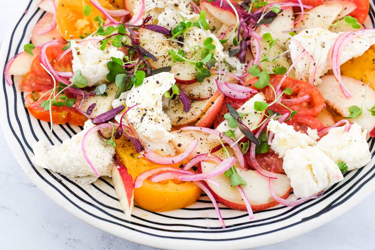 A salad of peaches, burrata cheese, tomato, red onion and micro greens sits on a marble countertop