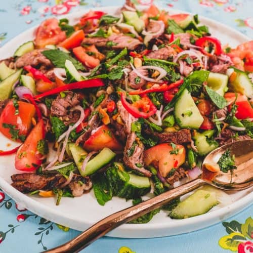 Thinly sliced ribeye steak is served in a salad of mint, cilantro shallots and spring onion in a spicy chilli dressing. Roasted peanuts are scattered over the salad. It sits non a bright blue floral plastic tablecloth like the ones in Thai markets.