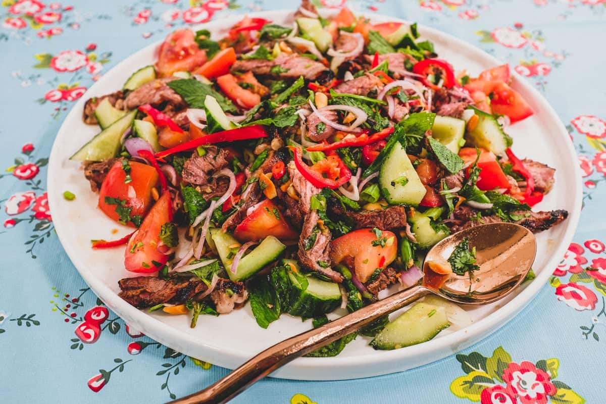 Thinly sliced ribeye steak is served in a salad of mint, cilantro shallots and spring onion in a spicy chilli dressing. Roasted peanuts are scattered over the salad. It sits non a bright blue floral plastic tablecloth like the ones in Thai markets.