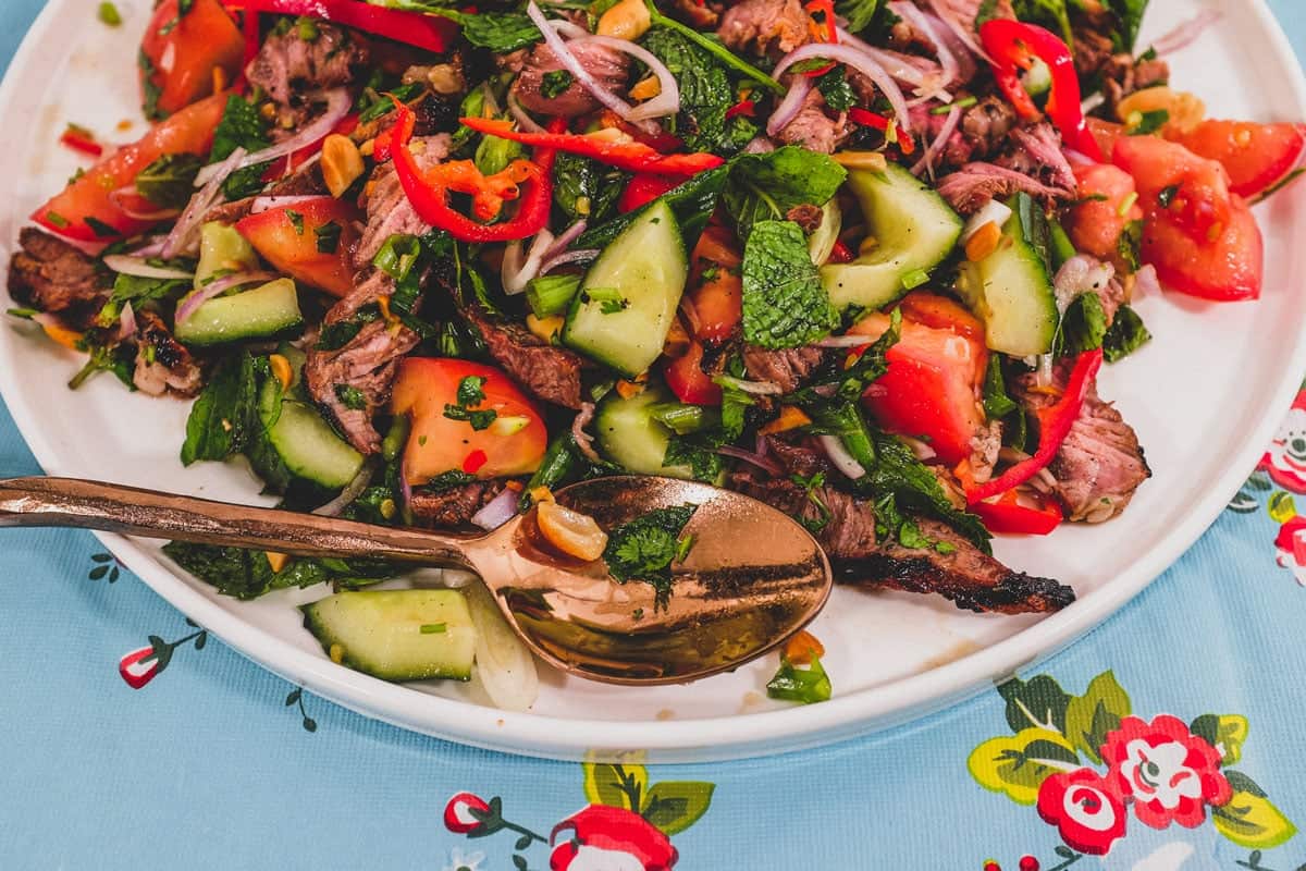 Thinly sliced ribeye steak is served in a salad of mint, cilantro shallots and spring onion in a spicy chilli dressing. Roasted peanuts are scattered over the salad. It sits non a bright blue floral plastic tablecloth like the ones in Thai markets.