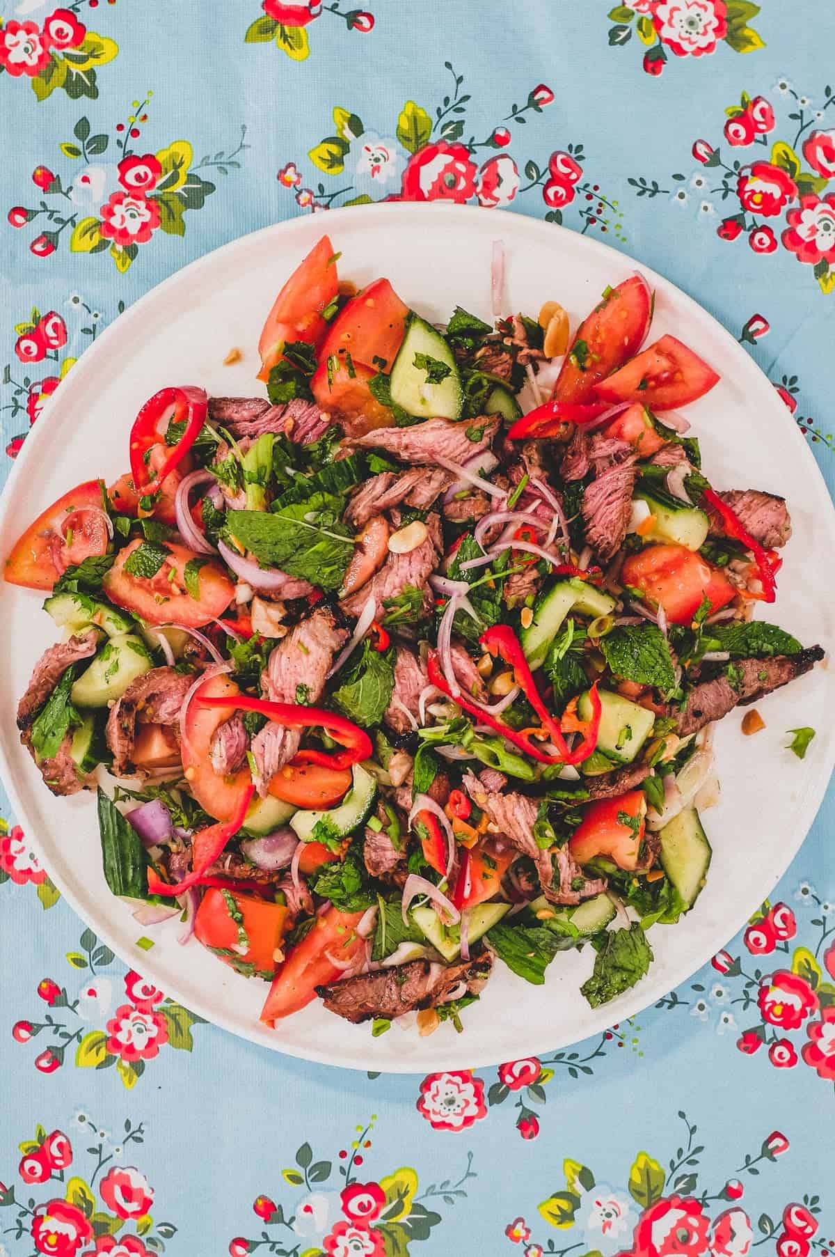 Thinly sliced ribeye steak is served in a salad of mint, cilantro shallots and spring onion in a spicy chilli dressing. Roasted peanuts are scattered over the salad. It sits non a bright blue floral plastic tablecloth like the ones in Thai markets.