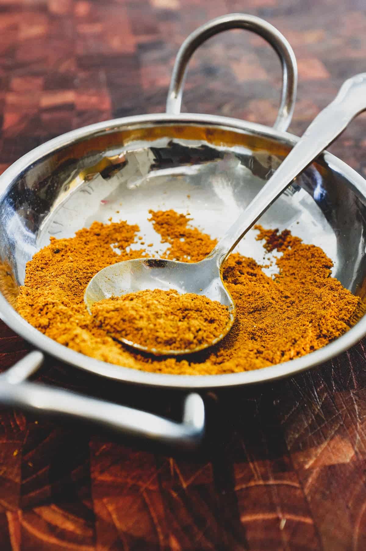 Ground spices combined with turmeric form a yellow spice powder called Hawaij, shown here in a silver bowl with spoon on an Arabic patterned background.