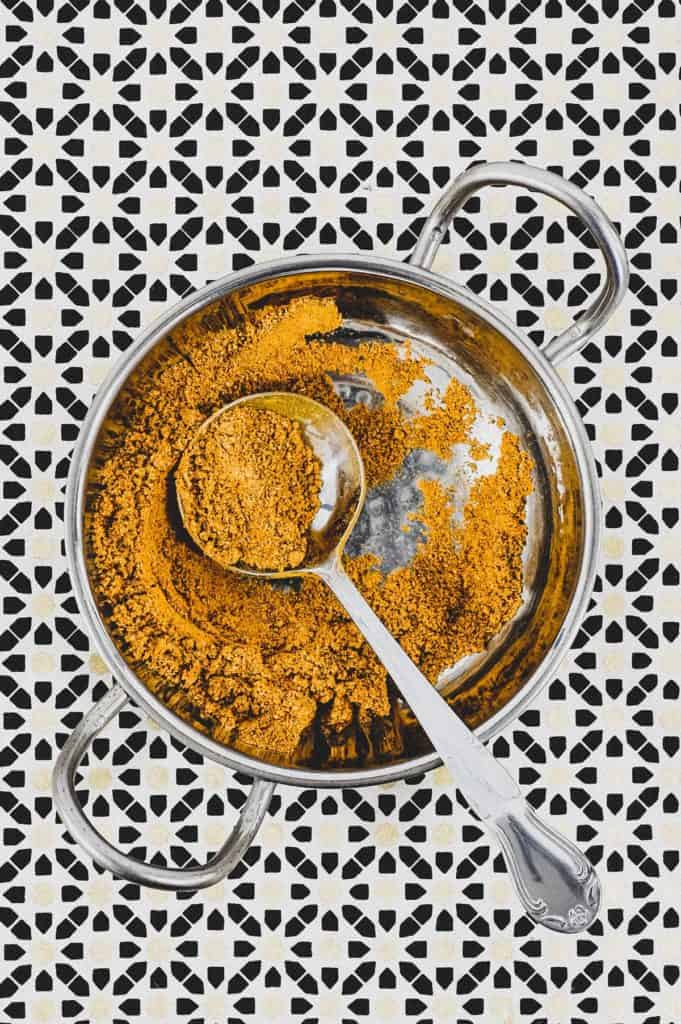 Ground spices combined with turmeric form a yellow spice powder called Hawaij, shown here in a silver bowl with spoon on an Arabic patterned background.