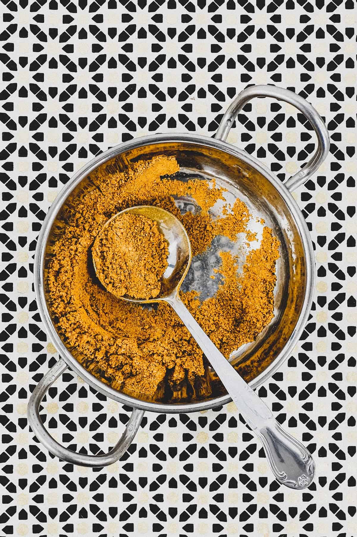 Ground spices combined with turmeric form a yellow spice powder called Hawaij, shown here in a silver bowl with spoon on an Arabic patterned background.