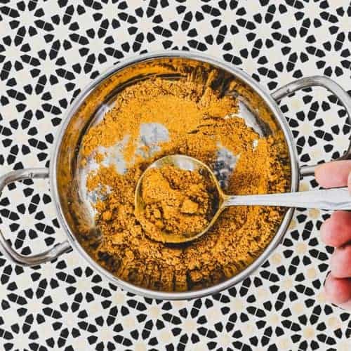 Ground spices combined with turmeric form a yellow spice powder called Hawaij, shown here in a silver bowl with spoon on an Arabic patterned background.