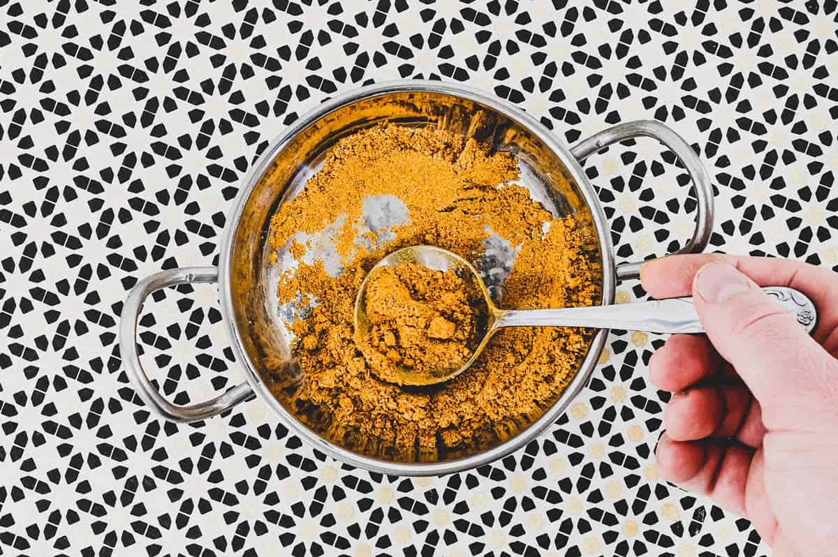 Ground spices combined with turmeric form a yellow spice powder called Hawaij, shown here in a silver bowl with spoon on an Arabic patterned background.