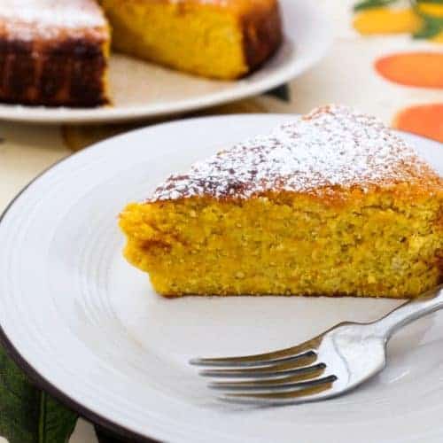 A slice of flourless orange cake sits on a plate with a fork. The rest of the cake is blurred in the background.
