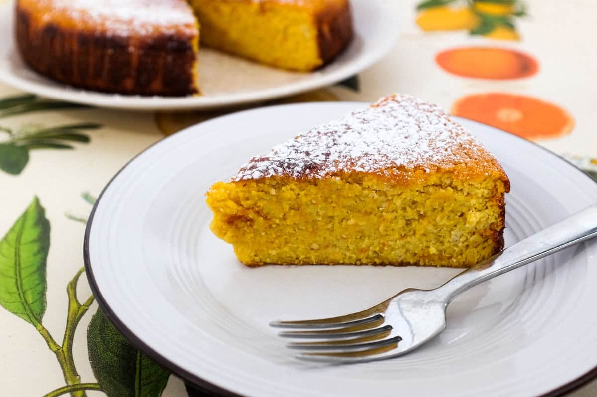 A slice of flourless orange cake sits on a plate with a fork. The rest of the cake is blurred in the background.