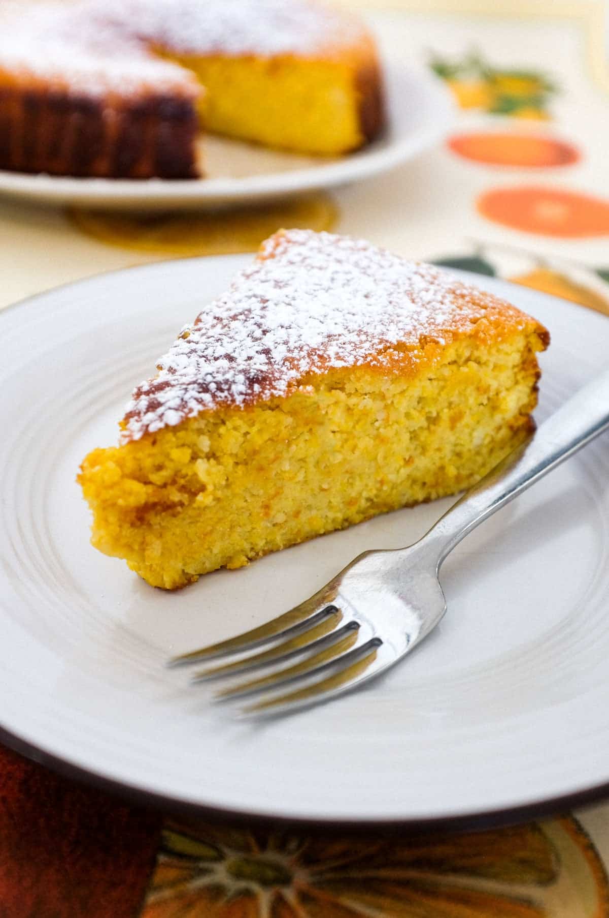 A slice of flourless orange cake sits on a plate with a fork. The rest of the cake is blurred in the background.