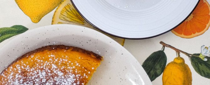 A flourless orange cake and slice of cake sit on plates on a colourful background of citrus fruit illustrations.