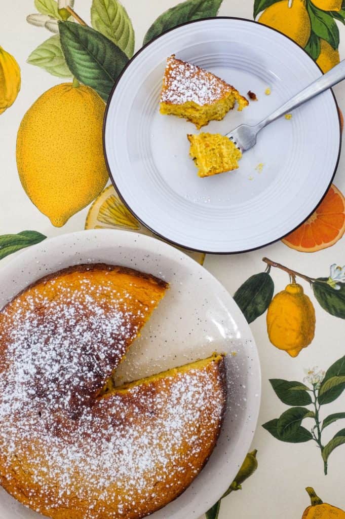 A flourless orange cake and slice of cake sit on plates on a colourful background of citrus fruit illustrations.
