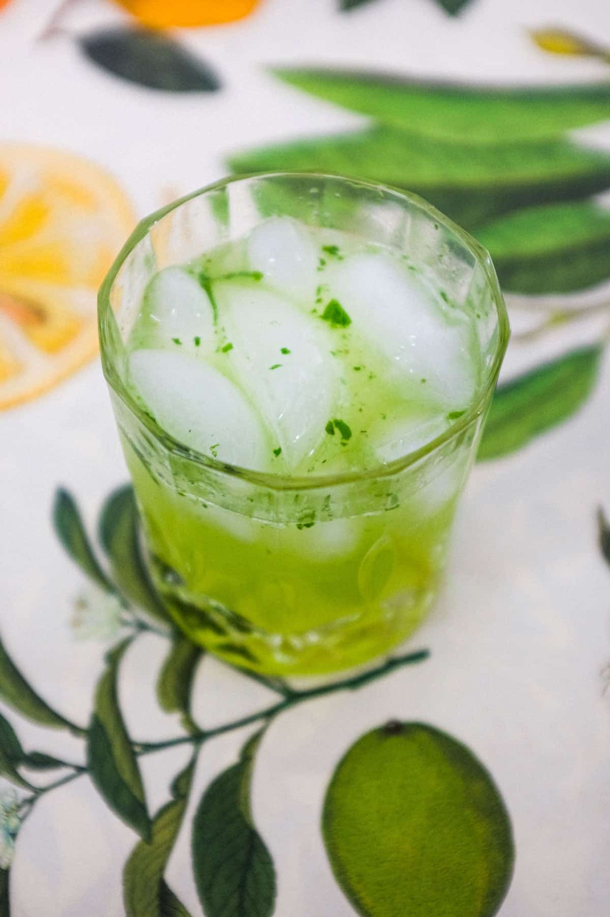 A green cocktail made from Gin and Thai Basil sits on a tablecloth of citrus fruits