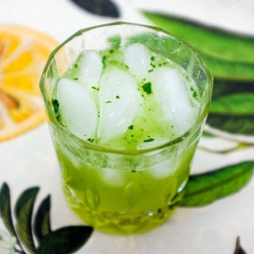 A green cocktail made from Gin and Thai Basil sits on a tablecloth of citrus fruits