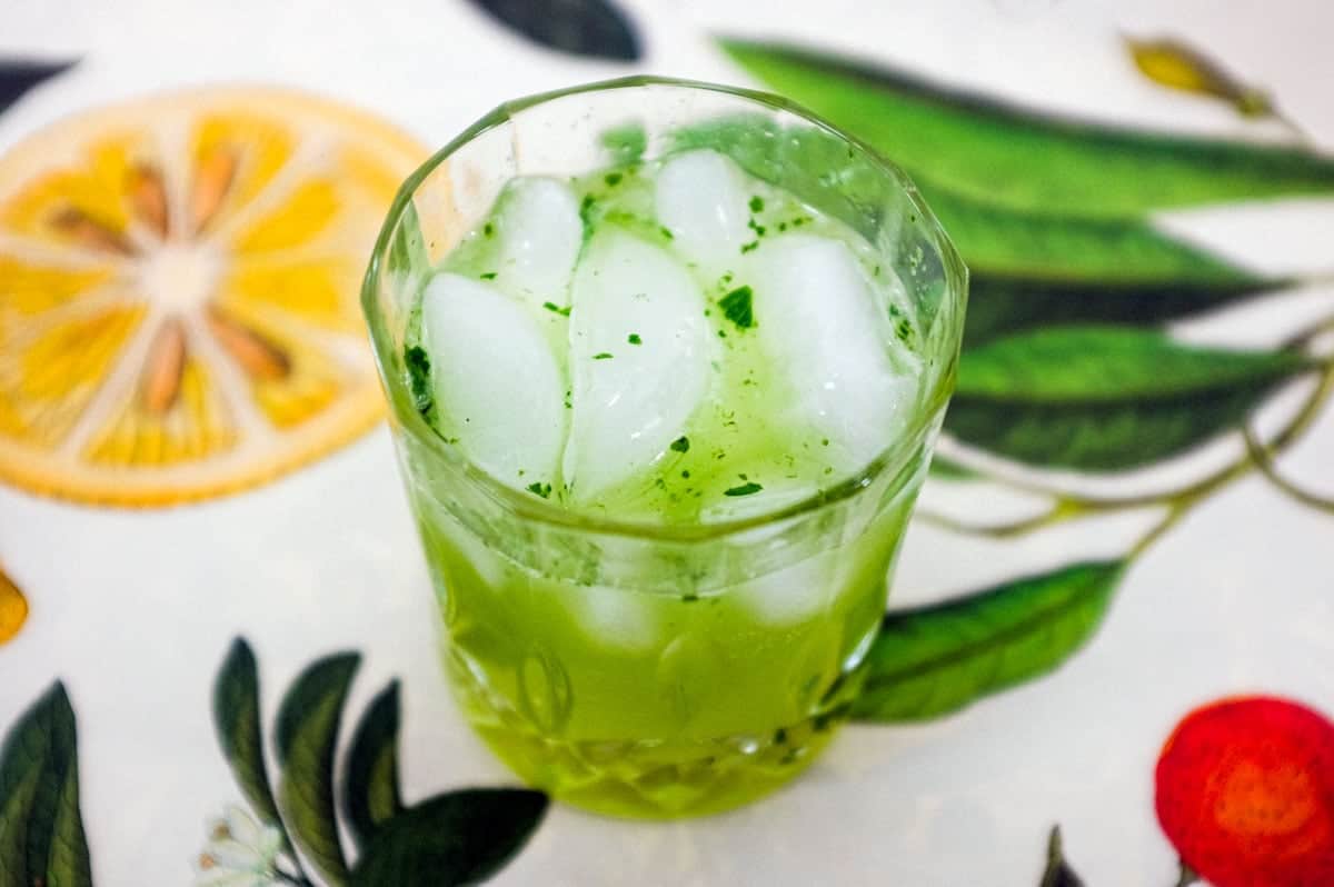 A green cocktail made from Gin and Thai Basil sits on a tablecloth of citrus fruits