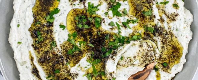 A plate of Haydari yoghurt dip, drizzled with olive oil and dried mint with a large copper spoon sit on a marble tabletop