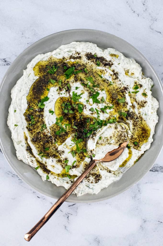 A plate of Haydari yoghurt dip, drizzled with olive oil and dried mint with a large copper spoon sit on a marble tabletop