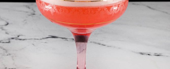 A pink Fizz cocktail with Campari and gin sits in a vintage Champagne glass