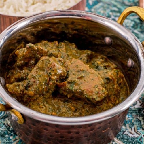 A serving of Saag Gosht in a copper pot with rice in the background and a large copper serving spoon sit on an antique silk carpet