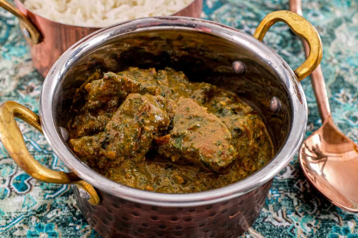 A serving of Saag Gosht in a copper pot with rice in the background and a large copper serving spoon sit on an antique silk carpet