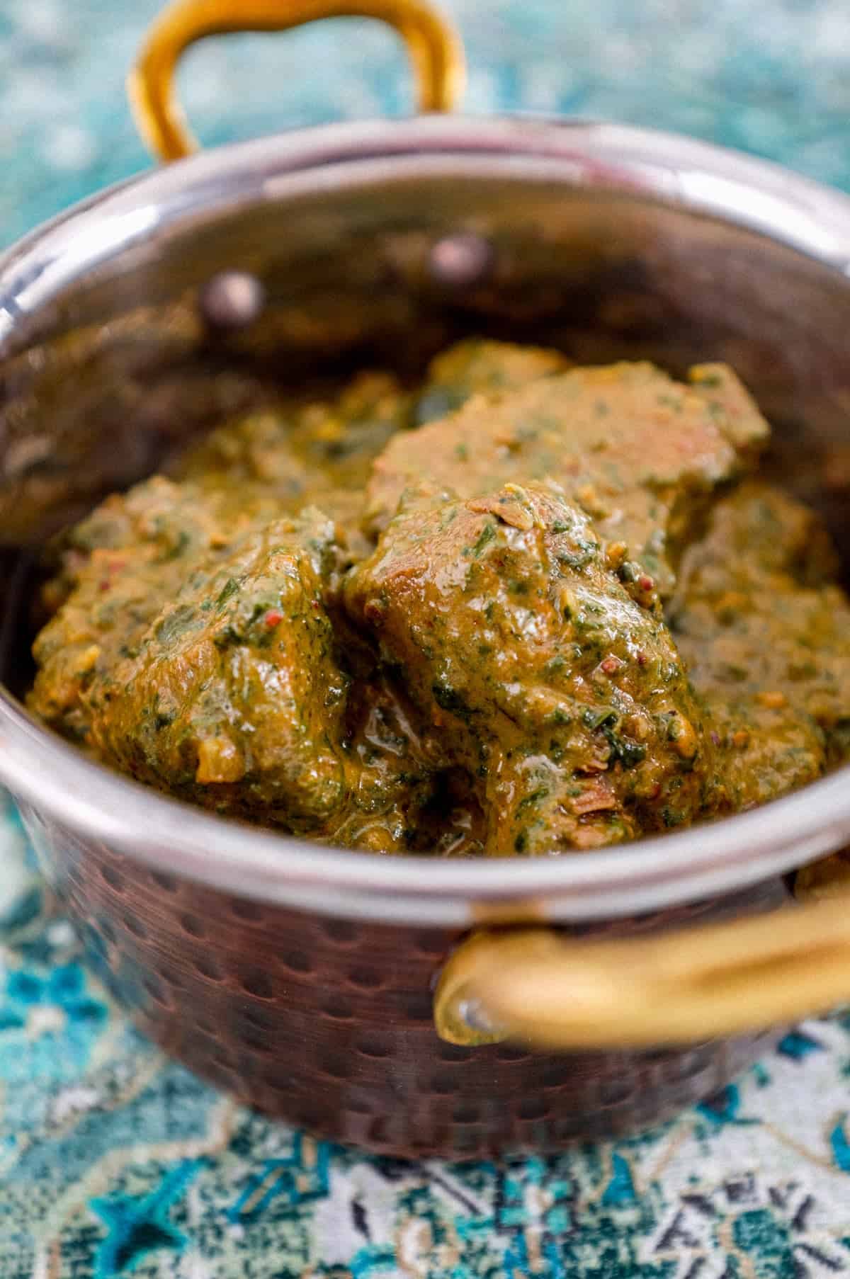 A serving of Saag Gosht in a copper pot with rice in the background and a large copper serving spoon sit on an antique silk carpet