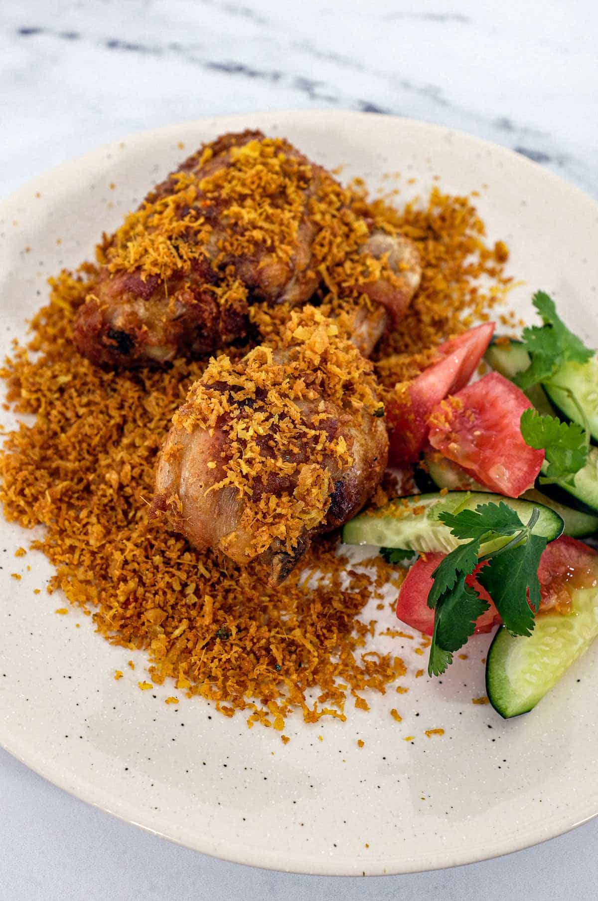 A platter of Ayam Serundeng fried chicken with crispy coconut with a side salad of cucumber and tomato