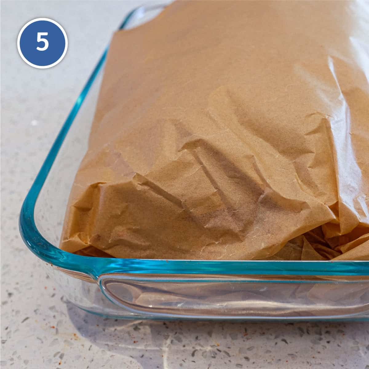 Pork butt wrapped in parchment paper in a glass baking dish