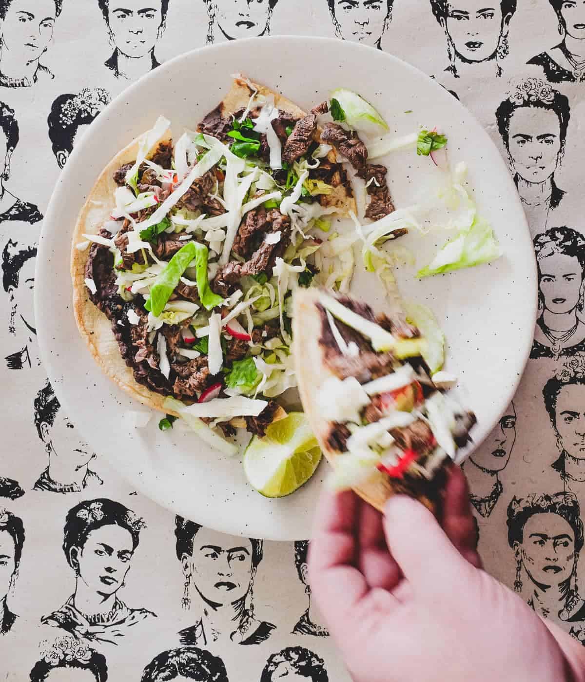 A plate with a Mexican tlayudas and wedge of lime. The background is a collage of sketches of Mexican artist Frida Kahlo