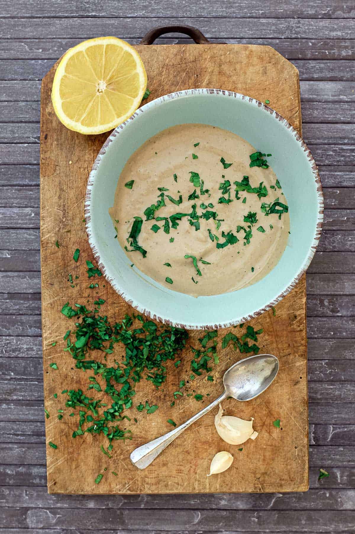 A bowl of anchovy sauce sits on a wooden board with lemon, garlic, parsley and a small spoon.