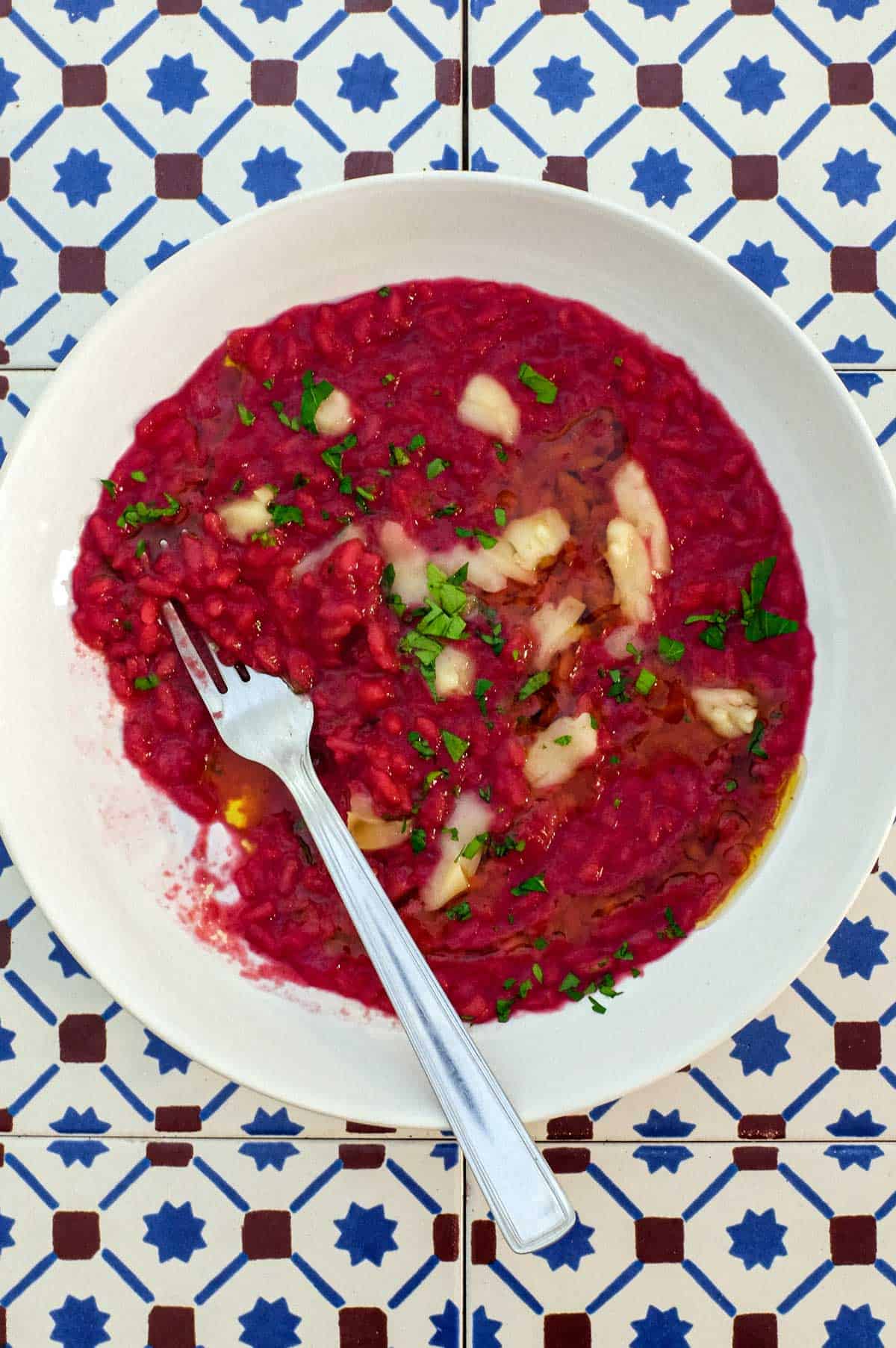 A bowl of beetroot risotto scattered with Taleggio cheese and fresh parsley on an Italian tiled background.