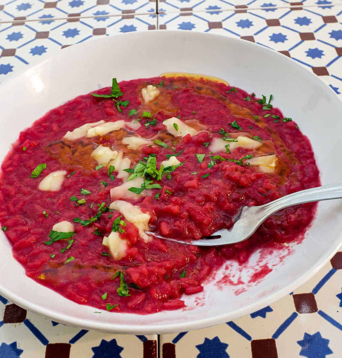 A bowl of beetroot risotto scattered with Taleggio cheese and fresh parsley on an Italian tiled background.