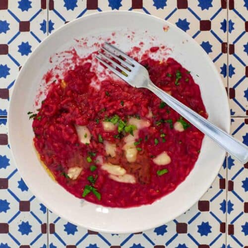 A bowl of beetroot risotto scattered with Taleggio cheese and fresh parsley on an Italian tiled background.