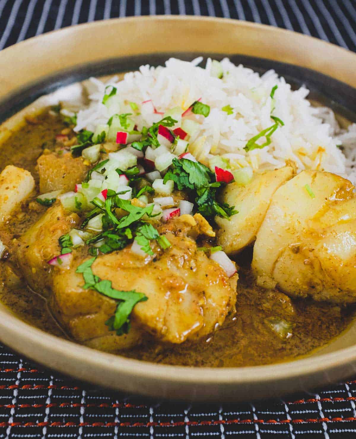 Bengali Fish Curry in a bowl in a black tabletop with rice and scattered with cilantro, radishes and celery