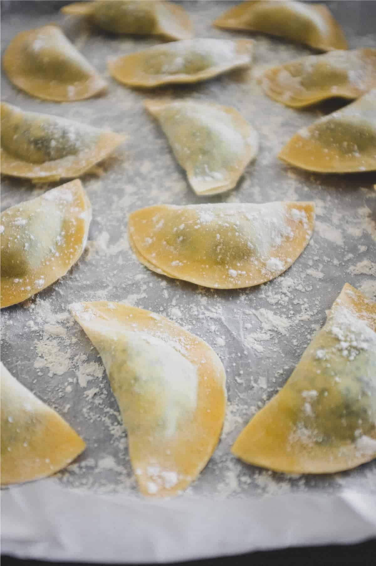 Freshly made mezzelune pasta half-moons on a baking tray lined with paper. The pasta is sprinkled with flour to stop them sticking.