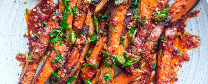 A plate of crisp and creamy sweet potato fries drizzled with a red sauce called Pilpelchuma. Fresh herbs and chillies are scattered over the potatoes too.