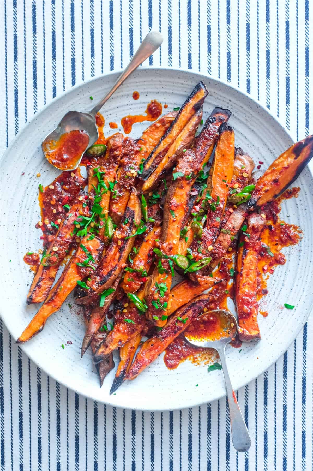 A plate of crisp and creamy sweet potato fries drizzled with a red sauce called Pilpelchuma. Fresh herbs and chillies are scattered over the potatoes too.