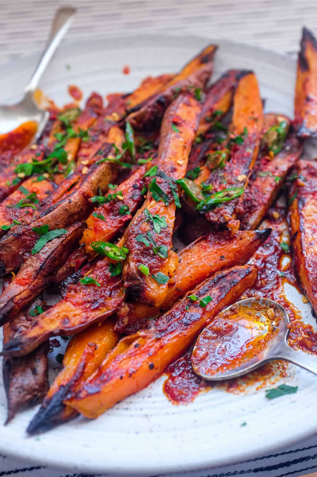 A plate of crisp and creamy sweet potato fries drizzled with a red sauce called Pilpelchuma. Fresh herbs and chillies are scattered over the potatoes too.