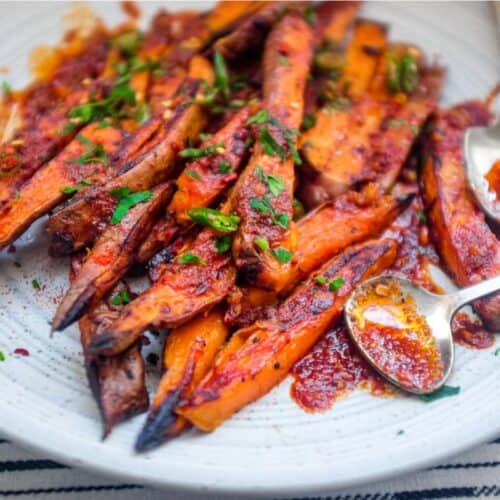 A plate of crisp and creamy sweet potato fries drizzled with a red sauce called Pilpelchuma. Fresh herbs and chillies are scattered over the potatoes too.