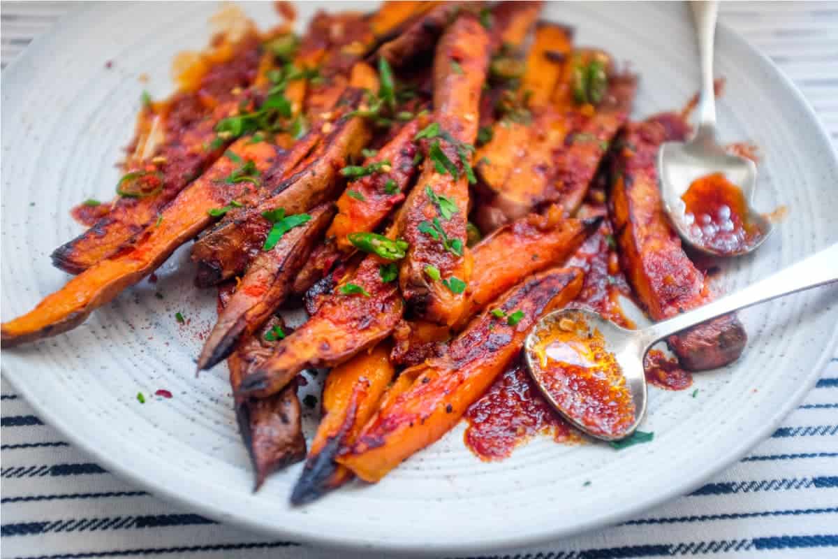 A plate of crisp and creamy sweet potato fries drizzled with a red sauce called Pilpelchuma. Fresh herbs and chillies are scattered over the potatoes too.