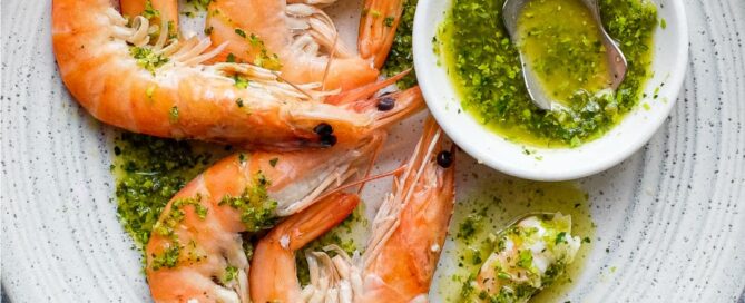 A platter of King Prawns with a small bowl of Italian Salmoriglio sauce, a herb and lemon dressing. A small spoon sits in the bowl.