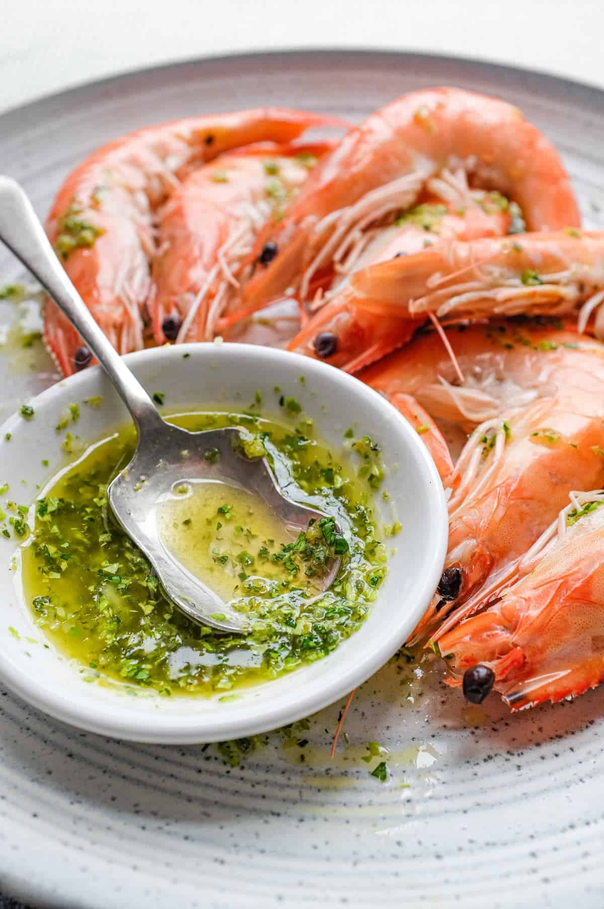 A platter of King Prawns with a small bowl of Italian Salmoriglio sauce, a herb and lemon dressing. A small spoon sits in the bowl.