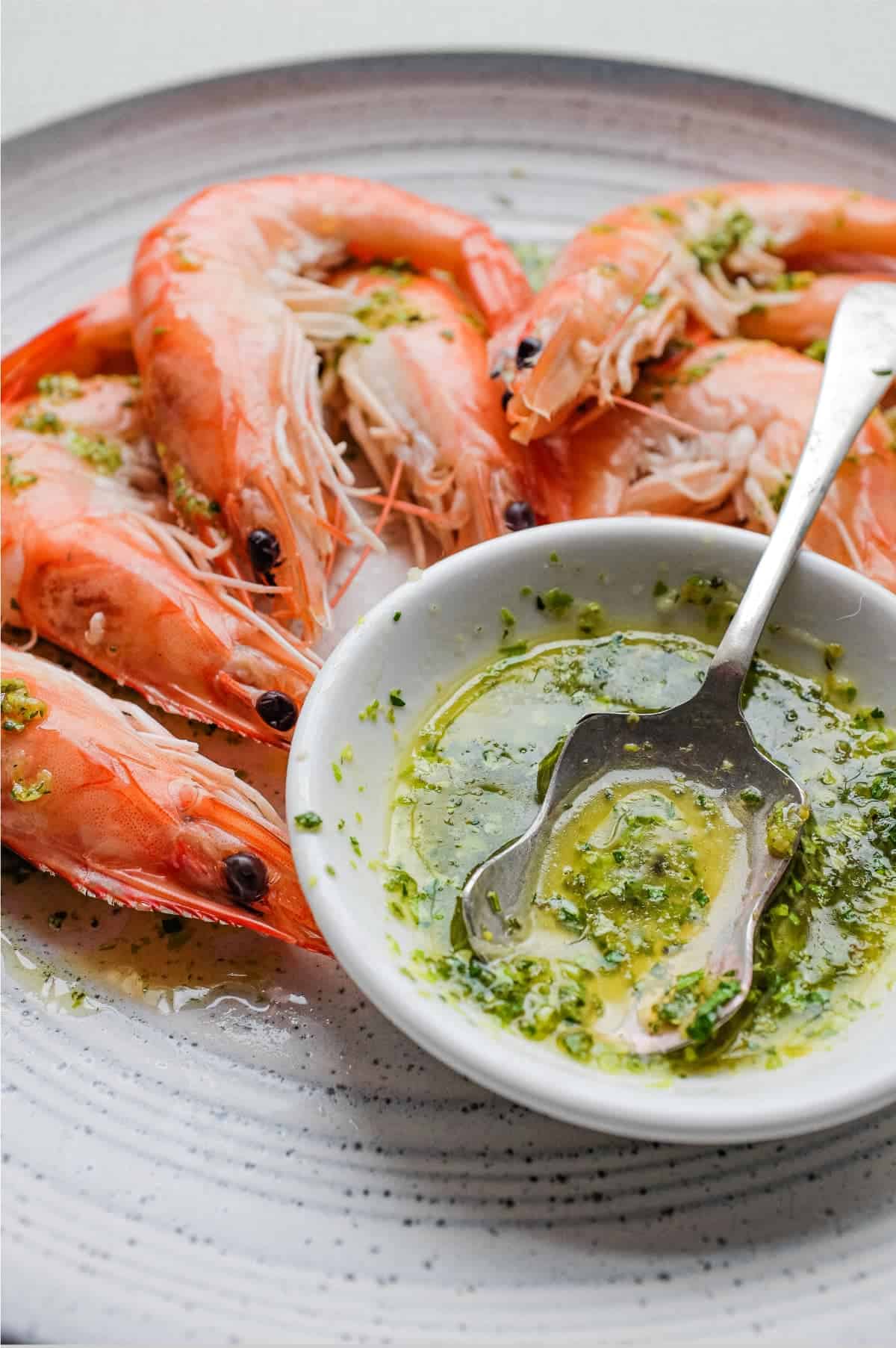 A platter of King Prawns with a small bowl of Italian Salmoriglio sauce, a herb and lemon dressing. A small spoon sits in the bowl.