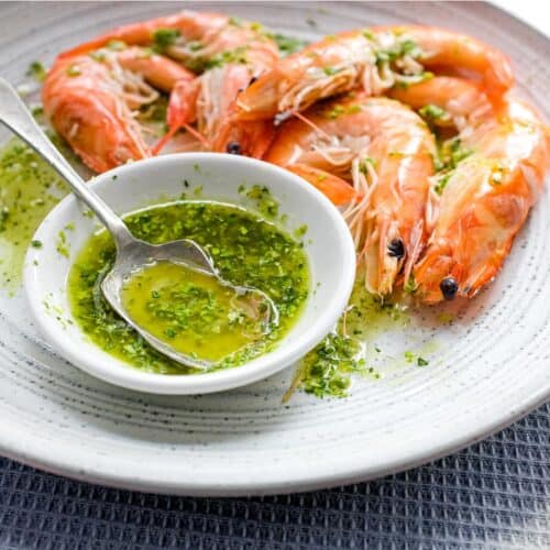 A platter of King Prawns with a small bowl of Italian Salmoriglio sauce, a herb and lemon dressing. A small spoon sits in the bowl.