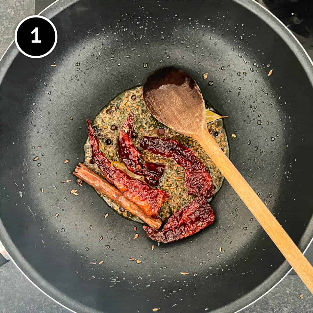 Frying whole spices and dried chillies in oil in a pan with a wooden spoon.