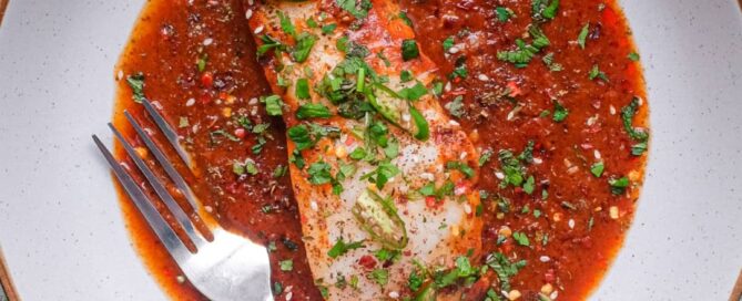 A fish fillet in a tomato Chraime sauce garnished with herbs and spices. A fork sits next to the fish