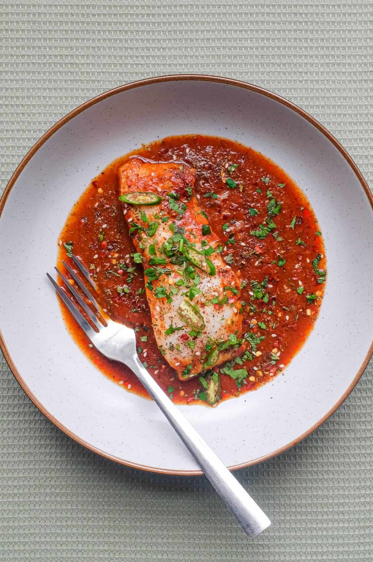 A fish fillet in a tomato Chraime sauce garnished with herbs and spices. A fork sits next to the fish