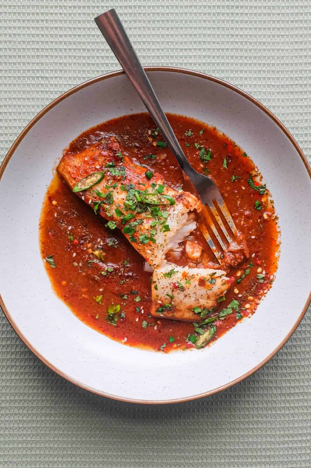 A fish fillet in a tomato Chraime sauce garnished with herbs and spices. A fork sits next to the fish