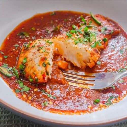 A fish fillet in a tomato Chraime sauce garnished with herbs and spices. A fork sits next to the fish