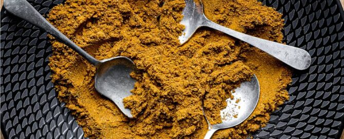 A black bowl of Madras Curry Powder on a wooden surface with three small spoons.