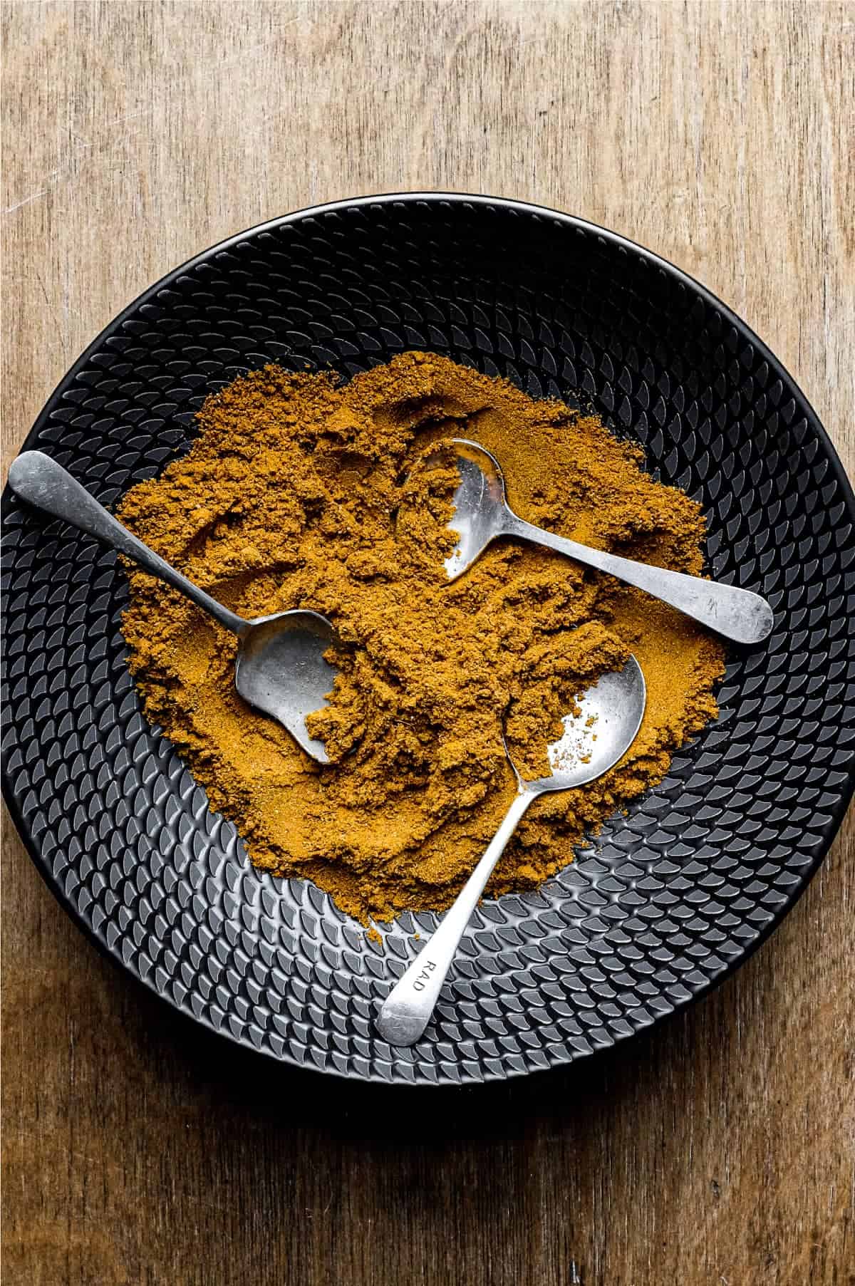 A black bowl of Madras Curry Powder on a wooden surface with three small spoons.